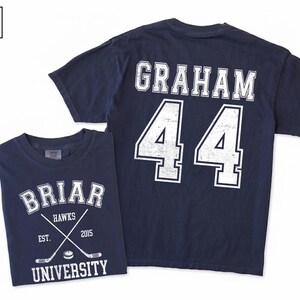 Puede incluir: Una camiseta azul marino de manga corta con un gr&aacute;fico blanco en la parte delantera que dice BRIAR UNIVERSITY HAWKS EST. 2015 con palos de hockey cruzados y un disco. La parte trasera muestra el nombre GRAHAM y el n&uacute;mero 44 en letras blancas.
