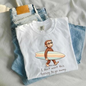 May include: White t-shirt with a cartoon monkey wearing sunglasses and carrying a surfboard. The text "I don't want this feeling to go away" is printed below the graphic. Light blue denim jeans are folded next to the shirt.