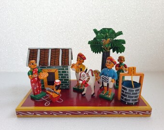 May include: A colorful wooden figurine set depicting a traditional Indian village scene. The set includes a small house, a palm tree, a well, a horse, and five figures. The figures are painted in bright colors and are wearing traditional clothing.