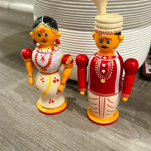May include: Two wooden dolls, a couple, stand side-by-side. The woman doll wears a white sari with red and yellow accents. The man doll wears a red and white outfit with a turban. Both dolls have painted faces and red arms.