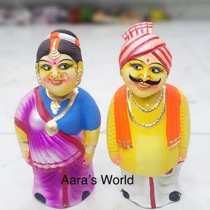 May include: Two colorful, handcrafted figurines of a man and woman. The woman wears a blue blouse and a purple sari. The man wears a yellow shirt, orange vest, and a turban. The text "Aara's World" is visible at the bottom.
