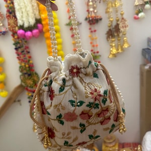 May include: A white drawstring bag with a floral pattern and gold accents. The bag has a pearl handle and is decorated with red, pink, and green flowers.