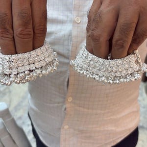 May include: Two silver anklets are held up, showcasing their intricate design. Each anklet features multiple rows of small, sparkling embellishments and dangling bells. The anklets are displayed against a light-colored background.