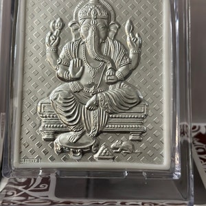 May include: A silver-colored, embossed, framed image of the Hindu deity Ganesha. The image is in a clear plastic frame with a decorative background.