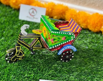 May include: A hand-painted wooden toy rickshaw with a green body, red and blue striped seat, and black wheels. The rickshaw is decorated with flowers and other colorful designs.