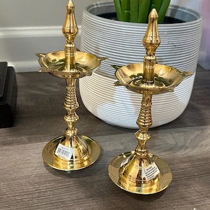 Puede incluir: Dos lámparas de aceite diya de metal dorado con un diseño decorativo. Las lámparas tienen una base redonda y un tallo alto y delgado con un pequeño cuenco redondo en la parte superior. Las lámparas están sobre una superficie de madera.