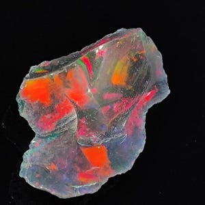 Puede incluir: Un ópalo bruto, translúcido e iridiscente con una mezcla de tonos rojos, naranjas, verdes y morados. El ópalo tiene una forma natural irregular.