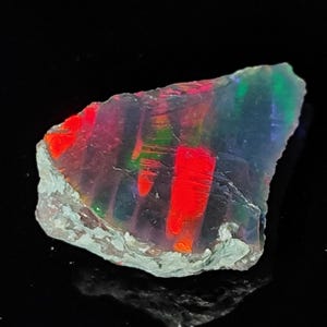 May include: A rough-cut opal gemstone with vibrant internal colors. The stone displays bands of red, green, and blue hues within a translucent matrix. The edges have a rough, natural texture, and the overall shape is irregular.