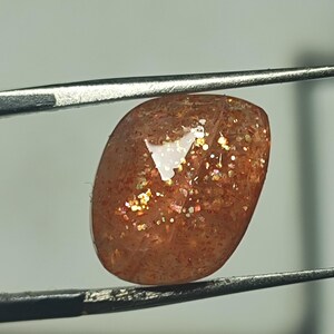 Op de afbeelding: Een zonnesteen cabochon edelsteen met een roodbruine kleur en een glinsterende, iriserende afwerking. De steen wordt vastgehouden door een pincet.