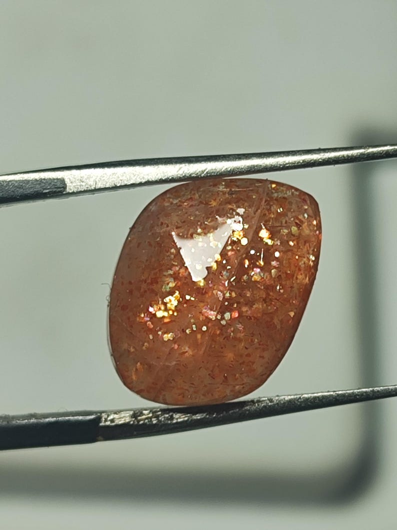 Op de afbeelding: Een zonnesteen cabochon edelsteen met een roodbruine kleur en een glinsterende, iriserende afwerking. De steen wordt vastgehouden door een pincet.