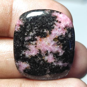 Puede incluir: Una piedra preciosa tallada en cabuj&oacute;n rosa y negra con un patr&oacute;n de remolino. La piedra preciosa est&aacute; pulida y tiene una superficie lisa.