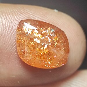 Op de afbeelding: Een sunstone edelsteen met een rood-oranje kleur en een glinsterende, iriserende afwerking. De steen is geslepen in een traanvorm en heeft een glad, gepolijst oppervlak.