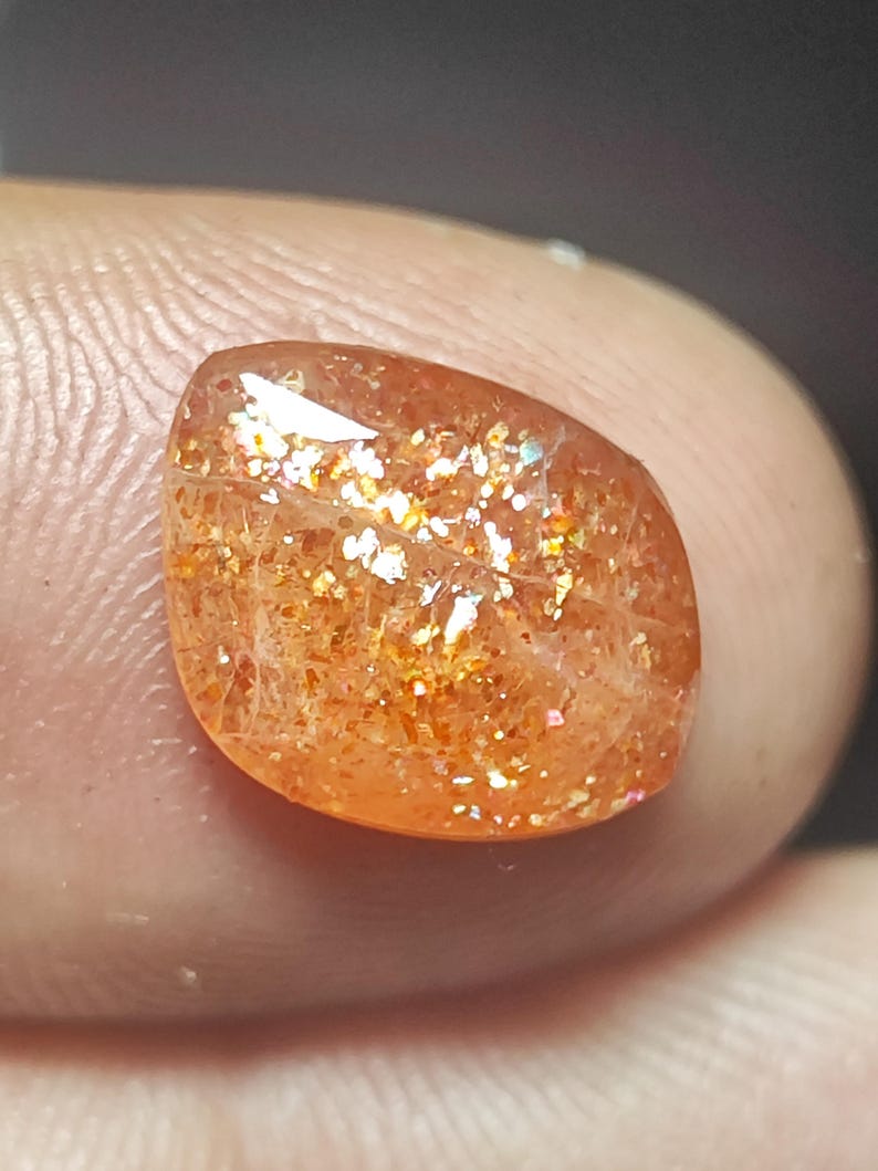 Op de afbeelding: Een sunstone edelsteen met een rood-oranje kleur en een glinsterende, iriserende afwerking. De steen is geslepen in een traanvorm en heeft een glad, gepolijst oppervlak.