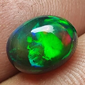 Könnte beinhalten: Ein polierter, ovaler schwarzer Opal-Edelstein. Der Opal zeigt lebendige interne Farben, darunter leuchtend grüne, blaue und rote Blitze. Der Stein hat eine glänzende Oberfläche und wird in einer Hand gehalten, was seine natürliche Schönheit zeigt.