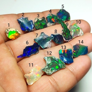 Op de afbeelding: Een verzameling ruwe opaal edelstenen in verschillende vormen en maten. De opalen vertonen een scala aan kleuren, waaronder blauw, groen, oranje en geel. De edelstenen worden in een hand gehouden, wat hun natuurlijke, ongepolijste staat laat zien.