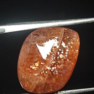Op de afbeelding: Een zonnesteen cabochon edelsteen met een roodachtig-oranje kleur en een glinsterend, iriserend effect. De steen is geslepen in een ovale vorm en heeft een glad, gepolijst oppervlak.