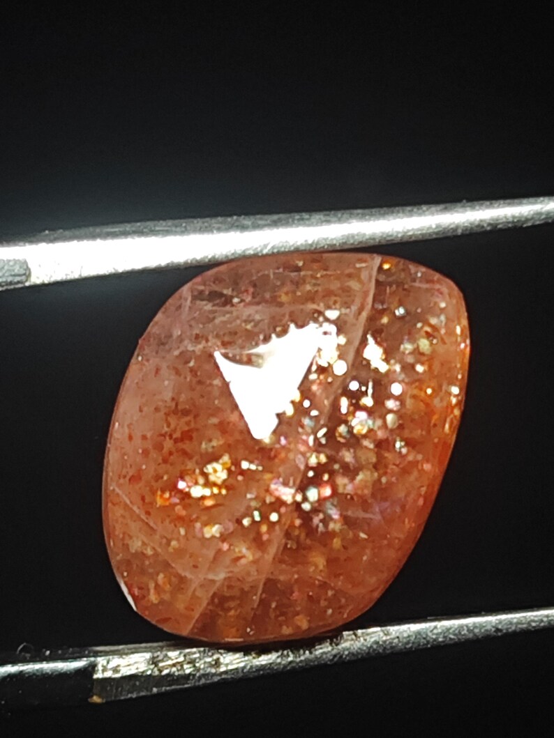 Op de afbeelding: Een zonnesteen cabochon edelsteen met een roodachtig-oranje kleur en een glinsterend, iriserend effect. De steen is geslepen in een ovale vorm en heeft een glad, gepolijst oppervlak.