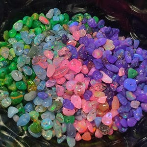 Könnte beinhalten: Eine Sammlung farbenfroher Opal-Edelsteine in Grün-, Blau-, Rosa- und Lilatönen. Die Opale sind kleine, unregelmäßige Formen und scheinen poliert zu sein. Die Edelsteine sind in einer Schüssel angeordnet und bilden eine lebendige Darstellung.