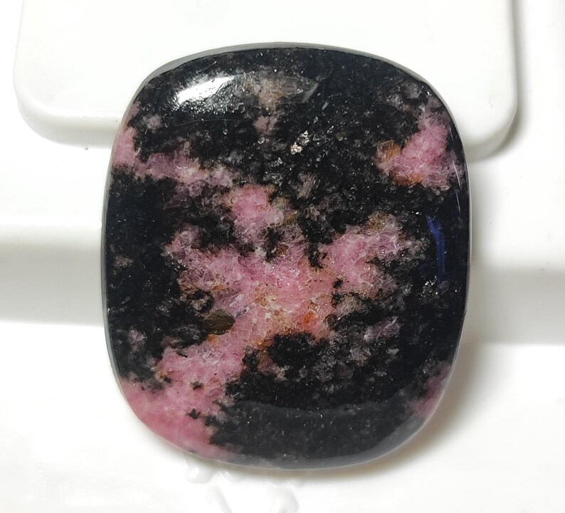 Puede incluir: Una piedra preciosa tallada en cabuj&oacute;n con un patr&oacute;n negro y rosa. La piedra tiene una superficie lisa y pulida y es un material natural y org&aacute;nico.