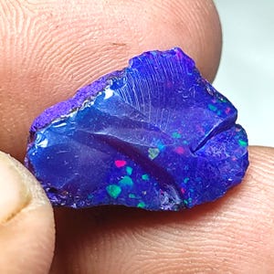 Op de afbeelding: Een ruw geslepen, gepolijste opaal edelsteen met een diepblauwe basiskleur. De opaal toont flitsen van roze, groen en blauw, met een paarse rand. Het oppervlak heeft zichtbare lijnen en texturen.