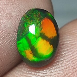 Könnte beinhalten: Ein oval geschliffener Edelstein mit lebendigen Farben. Der Stein zeigt eine Mischung aus Grün, Orange und Gelb auf dunklem Grund. Der Edelstein wird zwischen Fingern gehalten, wodurch seine reflektierende Oberfläche und die komplexen Farbmuster hervorgehoben werden.