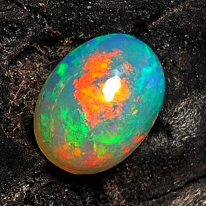 Könnte beinhalten: Ovale Opal-Edelstein mit einer lebendigen Farbgebung. Der Opal hat eine durchscheinende Basis mit internen Blitzen in Orange, Rot, Grün und Blau. Der Edelstein ist vor einem dunklen, strukturierten Hintergrund platziert.