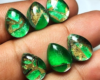 Puede incluir: Una colección de gemas en forma de lágrima. Cada gema presenta un color verde vibrante con vetas doradas. Las gemas se muestran sobre una superficie clara.