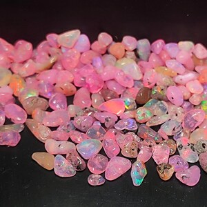 Könnte beinhalten: Eine Sammlung polierter, getrommelter Opal-Edelsteine in verschiedenen Rosa-, Pfirsich- und schillernden Farbtönen. Die Opale sind kleine, unregelmäßige Formen und scheinen über eine dunkle Oberfläche verstreut zu sein.