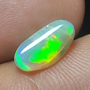 Könnte beinhalten: Ein ovaler Opal-Edelstein mit einer durchscheinenden, milchig-weißen Basis. Der Stein zeigt lebhafte grüne und gelbe Blitze mit blauen Farbtönen. Der Opal ist glatt und poliert und reflektiert das Licht über seine Oberfläche.