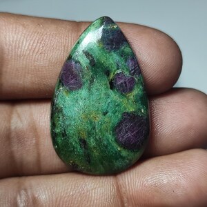 Op de afbeelding: Een groene en paarse edelsteen cabochon met een traanvorm. De edelsteen heeft een gevlekt patroon met donkerpaarse insluitsels.