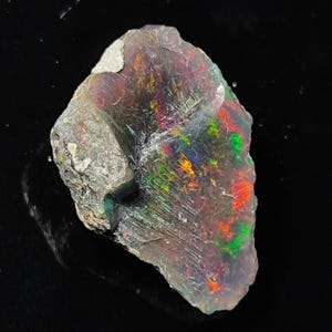 May include: A rough, gray, and white rock with a large, iridescent opal embedded in it. The opal displays a rainbow of colors, including red, orange, yellow, green, blue, and purple.