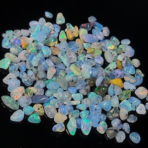 Könnte beinhalten: Eine Sammlung polierter Opal-Edelsteine in verschiedenen Formen und Größen. Die Opale zeigen eine Reihe von Farben, darunter Blau-, Grün-, Gelb- und schillernde Blitze. Die Edelsteine sind auf einer schwarzen Oberfläche verstreut.
