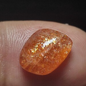 Op de afbeelding: Een zonnesteen cabochon met een roodachtig-oranje kleur en een glinsterende, iriserende afwerking. De steen is geslepen in een rechthoekige vorm en heeft een glad, gepolijst oppervlak.