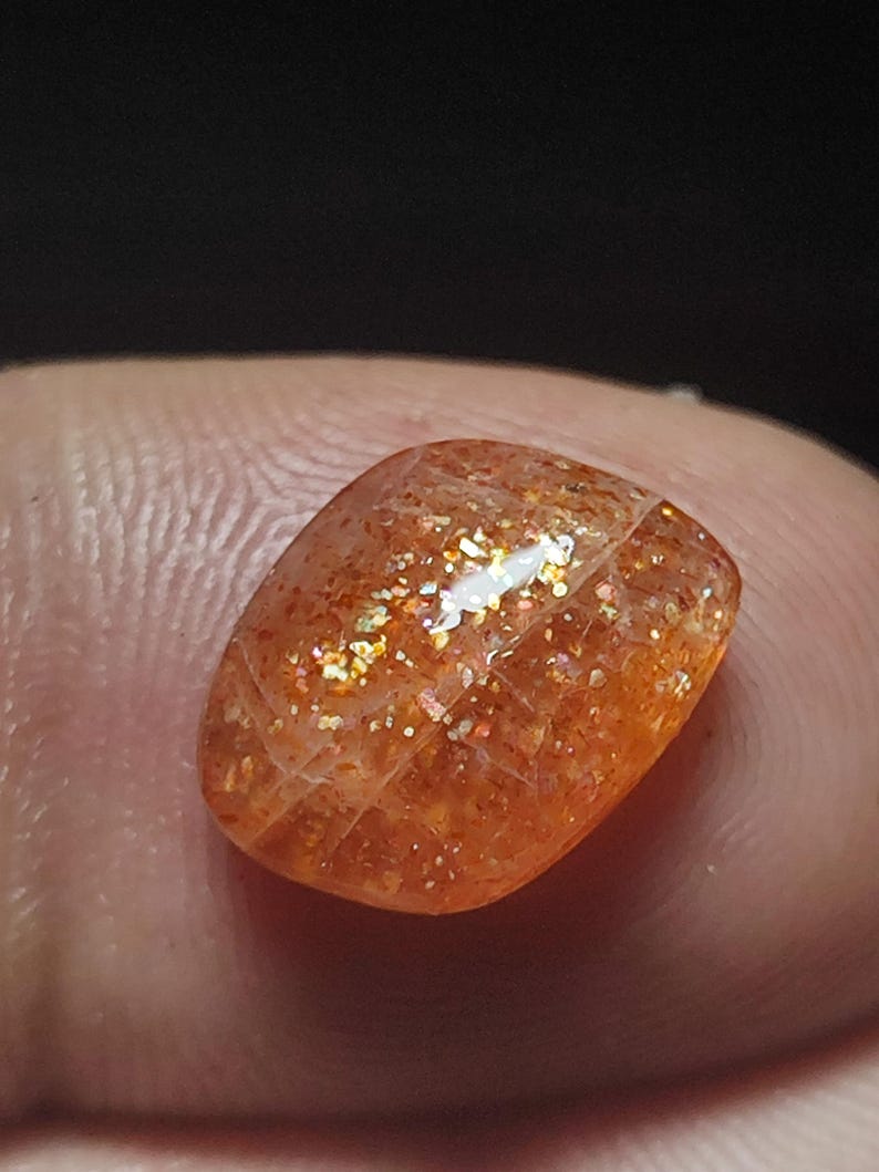 Op de afbeelding: Een zonnesteen cabochon met een roodachtig-oranje kleur en een glinsterende, iriserende afwerking. De steen is geslepen in een rechthoekige vorm en heeft een glad, gepolijst oppervlak.