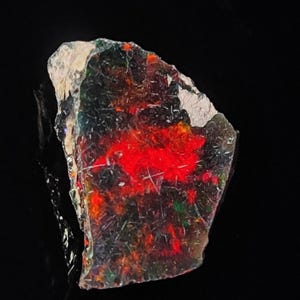 May include: A rough, irregular piece of red, green and black opal with a white edge. The opal has a strong red flash.