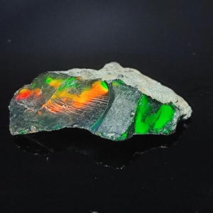 Könnte beinhalten: Ein rauer, grauer Stein mit einem grünen und orangefarbenen Opal, der darin eingebettet ist. Der Opal hat einen irisierenden Glanz und Farbschimmer.
