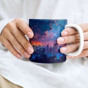 May include: A white ceramic mug with a colourful cityscape design. The mug features a nighttime cityscape with a purple, blue, and orange sky. The cityscape is silhouetted against the sky.