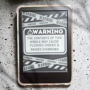 Op de afbeelding: Een zwart-wit waarschuwingsbord op een Kindle e-reader. Het bord luidt "WAARSCHUWING: De inhoud van deze Kindle kan leiden tot rode wangen en opgeheven wenkbrauwen."