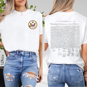 May include: White t-shirt featuring the US seal on the front and the Declaration of Independence on the back. The shirt is a classic crew neck style and is paired with blue jeans. The text on the back reads "In Congress, July 4, 1776."