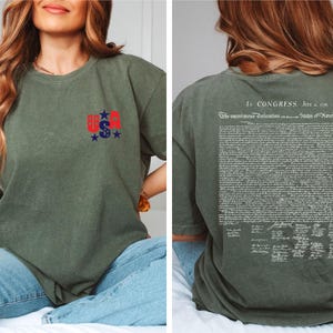 May include: Olive green t-shirt with a small "USA" graphic in red, white, and blue on the front. The back of the shirt features the text of the Declaration of Independence in white. The shirt is made of a soft material.