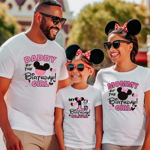 May include: A family of three wearing white t-shirts with pink and black Mickey Mouse designs. The shirts say "Daddy of the Birthday Girl", "Mommy of the Birthday Girl", and "My 1st Birthday Olivia".