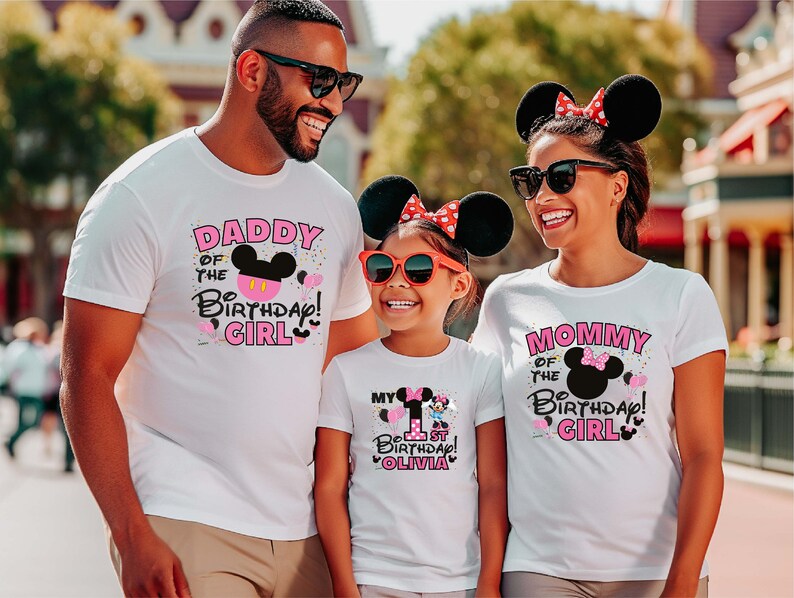 May include: A family of three wearing white t-shirts with pink and black Mickey Mouse designs. The shirts say "Daddy of the Birthday Girl", "Mommy of the Birthday Girl", and "My 1st Birthday Olivia".