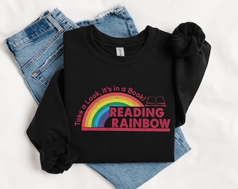 May include: Black sweatshirt with the text "Take a Look, it's in a Book! READING RAINBOW" in a rainbow design. The sweatshirt is folded on top of blue jeans.