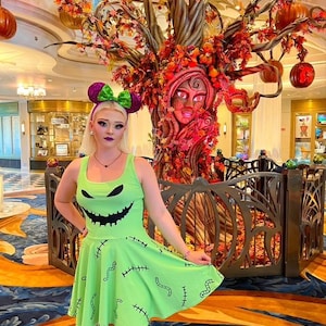 May include: A woman wearing a green dress with a black Jack Skellington face and stitches print. She is standing in front of a large, decorated tree with pumpkins.