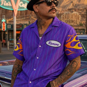 May include: A short-sleeved purple button-down shirt featuring thin vertical blue pinstripes, an oval patch on the chest with the name Ramone printed in black script, and orange and yellow flame graphics on the sleeves with a classic collar.