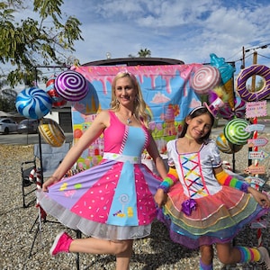 May include: Two people dressed in colorful costumes stand in front of a candy-themed backdrop. The person on the left is wearing a pink and blue dress with a white petticoat. The person on the right is wearing a white dress with rainbow trim and a purple hat. The backdrop features a castle, candy canes, and lollipops.