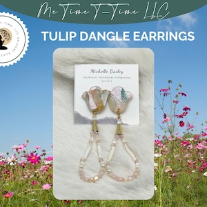Puede incluir: Un par de pendientes colgantes con un dise&ntilde;o de tulip&aacute;n rosa y verde con detalles dorados. Los pendientes est&aacute;n hechos de joyer&iacute;a ind&iacute;gena hecha a mano y cuelgan de una cadena de cuentas blancas.