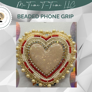 May include: A heart-shaped beaded phone grip. The grip features a central light pink stone, surrounded by red, white, and gold beads. The text "BEADED PHONE GRIP" is visible above the grip. The logo of "Me Time T-Time LLC" is at the top.