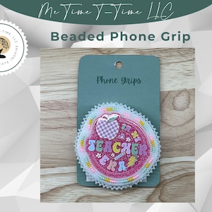 May include: A beaded phone grip with a pink and white design. The grip features the text "Teacher Era" with an apple graphic. The grip is attached to a green card that reads "Phone grips". The background is a wooden surface.