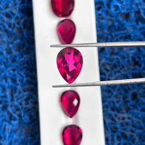 Puede incluir: Una fila de cinco piedras preciosas rosadas en forma de lágrima, facetadas, sobre un fondo blanco. Una piedra preciosa está siendo sostenida por unas pinzas.